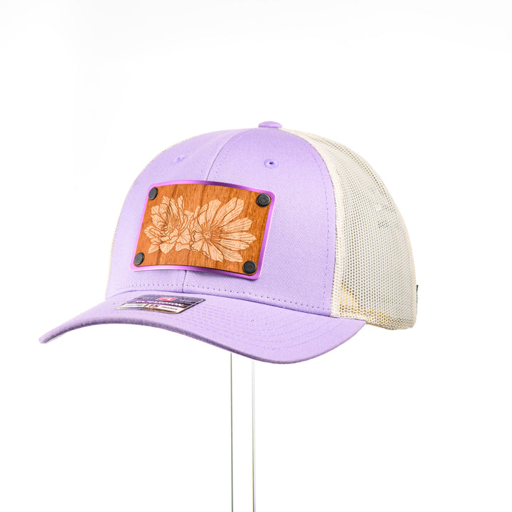 Cherry Wood Bitterroot Flower Patch Lilac & Cream Trucker Hat, featuring a real cherry wood veneer patch etched with a bitterroot flower, laminated to a light purple anodized aluminum plate and riveted to a lilac and cream trucker hat along—handmade in Montana, USA by Last Best Supply Co.