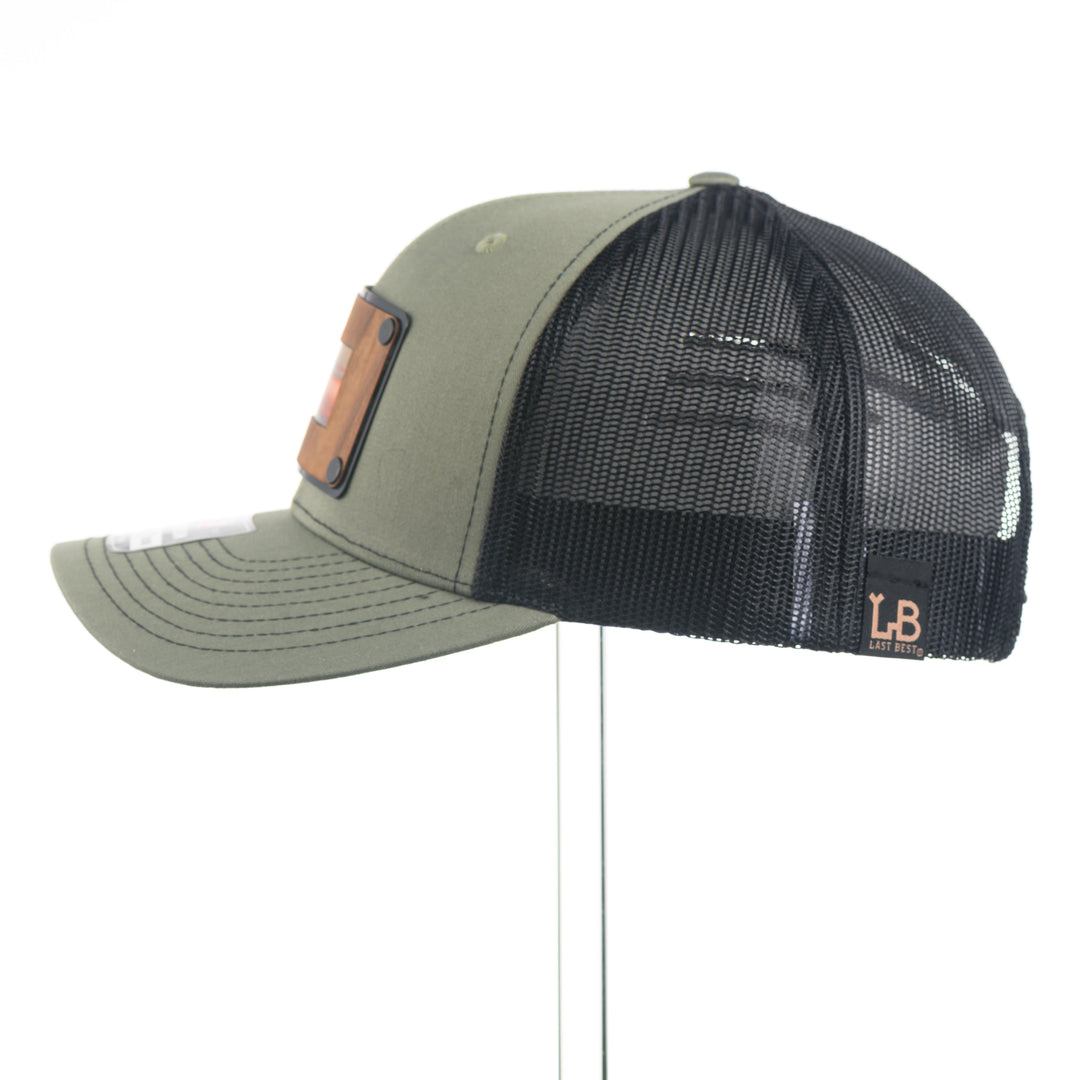 Side view of a Montana Walnut Wood with Sundae Copper Patch on Richardson Trucker Hat, featuring a rectangular, real walnut wood veneer patch with a precision-cut state of Montana silhouette cutout, laminated to a black anodized metal plate, and then riveted to an olive green Richardson 112 trucker hat with black mesh—handmade in Montana, USA by Last Best Supply Co.