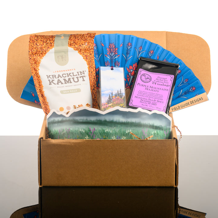 A Montana made gift box featuring a bag of Kracklin Kamut khorasan wheat snacks, a prairie fire/ (Indian) paintbrush hand fan, a watercolor bookmark, a bag of Montana Tea & Spice Co.'s Purple Mountain Majesty tea, and a large wildflower sticker wrap.