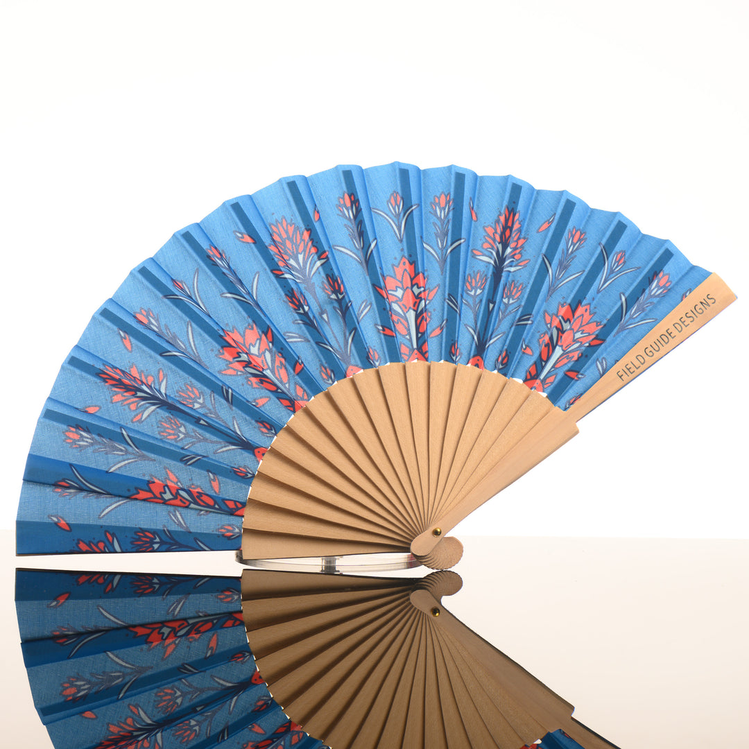 A Montana made artist hand fan featuring a blue background with hand drawn prairie fire/ (Indian) paintbrush flowers, by Fieldguide Design, featured in The Petals to Peaks Box by The Last Best Box.