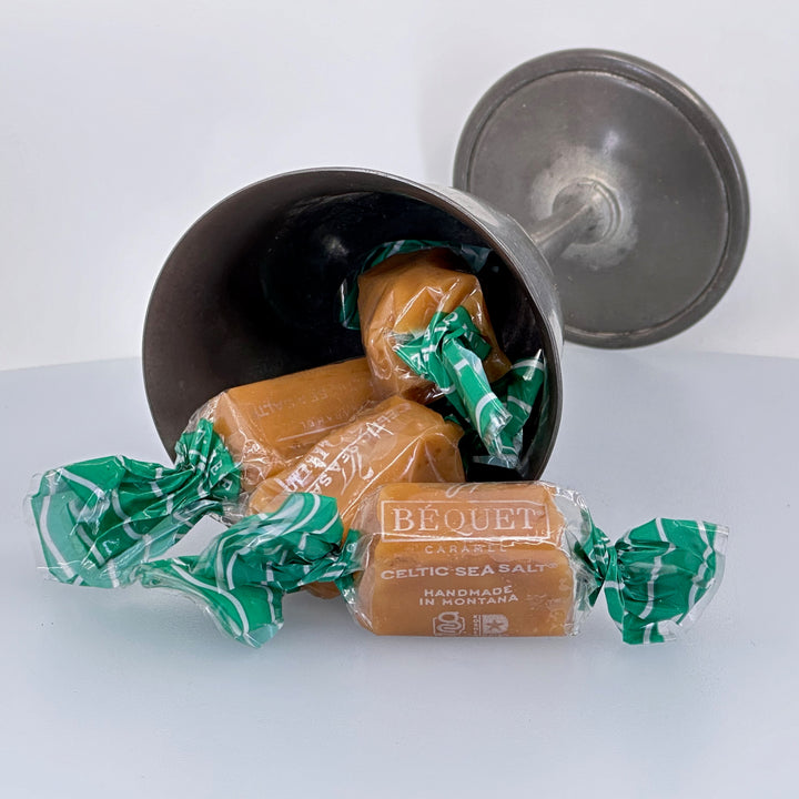 Bequet sea salt caramels in green and white wrappers on a white surface with a metal container, made in montana