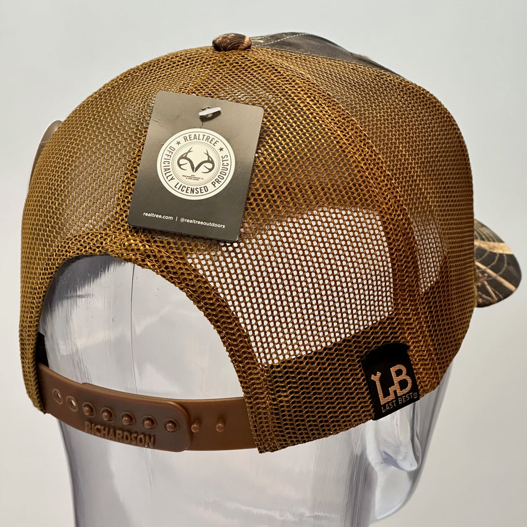 A back view of a made in Montana wood patch of an etched pheasant on a gold metal plate riveted with black rivets to a real tree camo Richardson trucker hat.