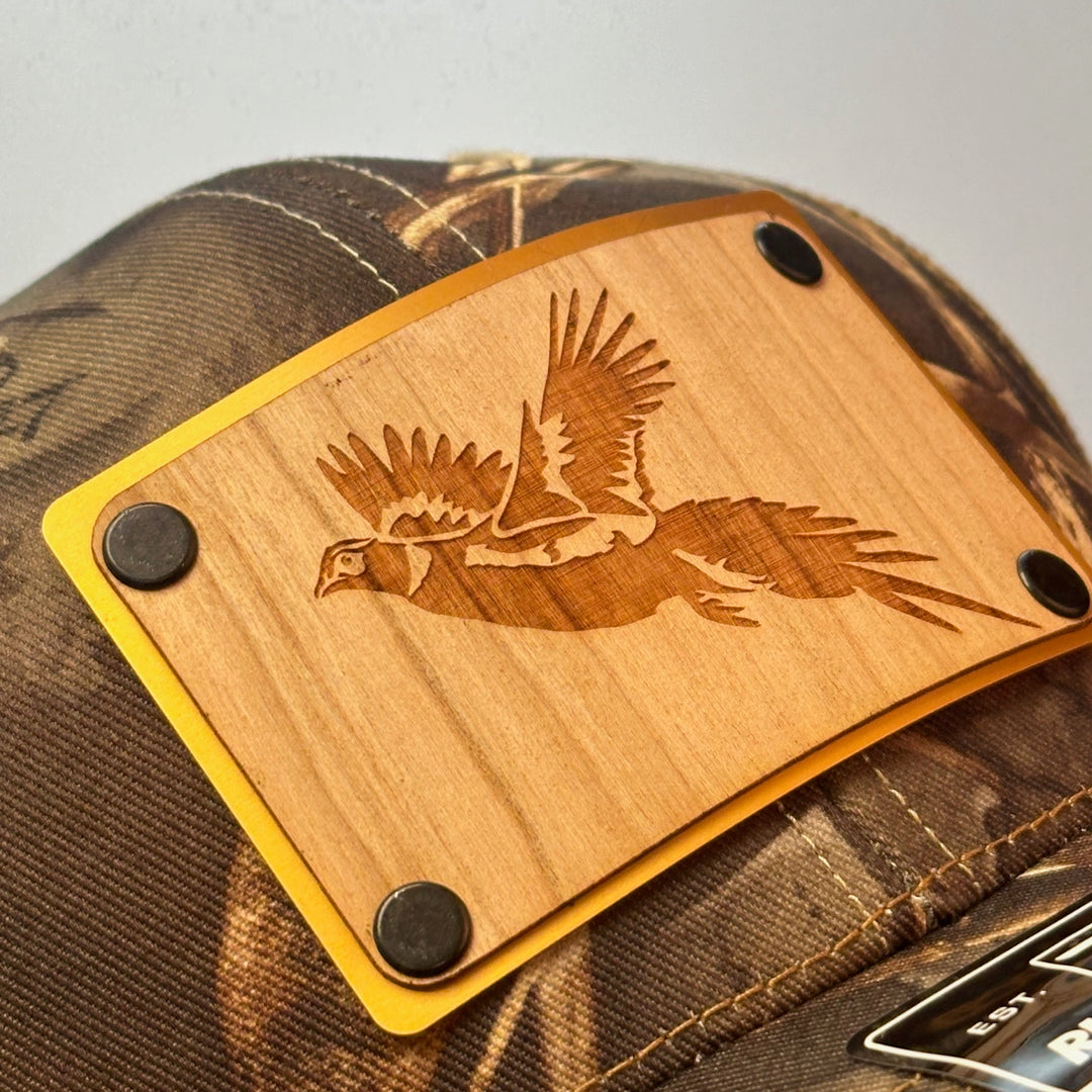 A close up of a made in Montana wood patch of an etched pheasant on a gold metal plate riveted with black rivets to a real tree camo Richardson trucker hat.