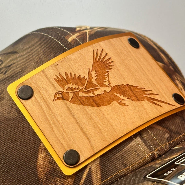 A close up of a made in Montana wood patch of an etched pheasant on a gold metal plate riveted with black rivets to a real tree camo Richardson trucker hat.