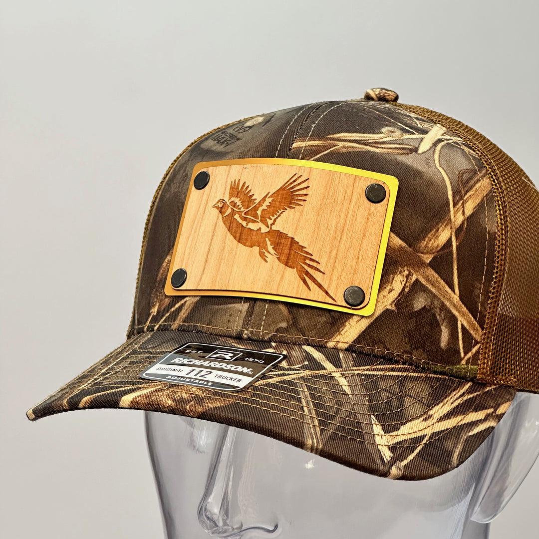 A left side view of a made in Montana wood patch of an etched pheasant on a gold metal plate riveted with black rivets to a real tree camo Richardson trucker hat.