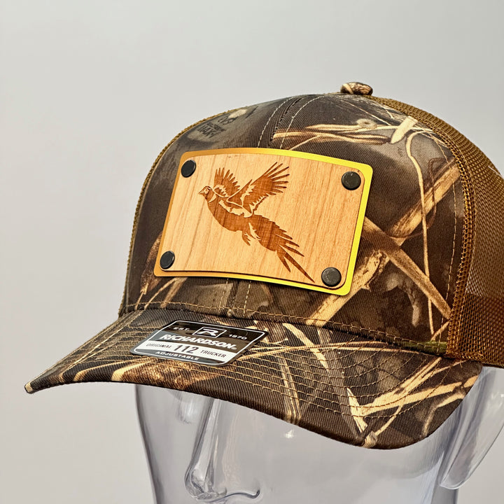 A left side view of a made in Montana wood patch of an etched pheasant on a gold metal plate riveted with black rivets to a real tree camo Richardson trucker hat.