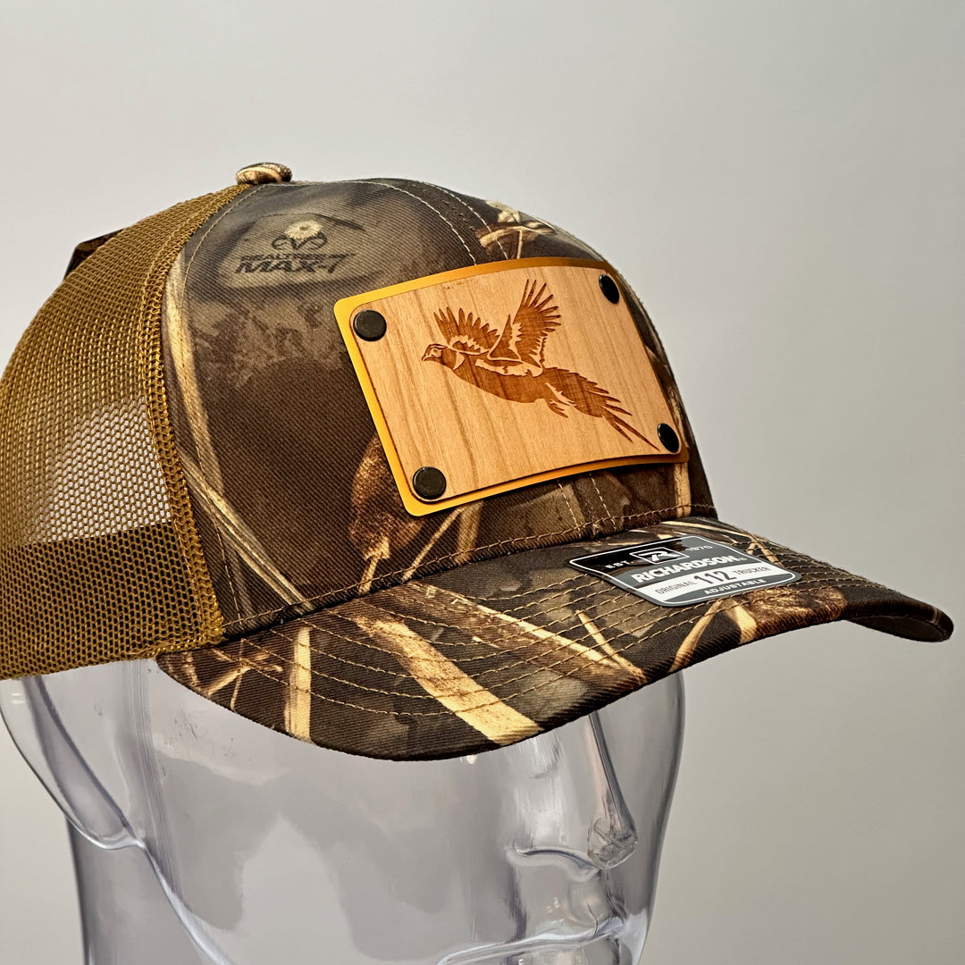 A right side view of a made in Montana wood patch of an etched pheasant on a gold metal plate riveted with black rivets to a real tree camo Richardson trucker hat.