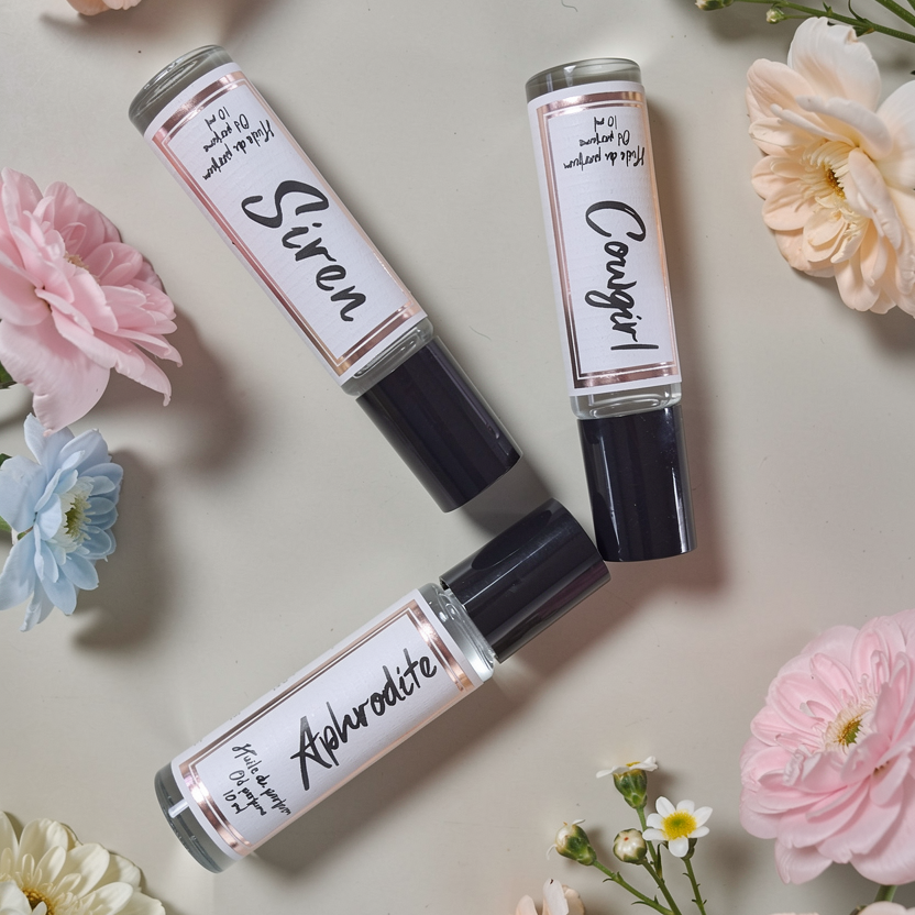 Three perfume roller tubes labeled 'Siren', 'Candied', and 'Aphrodite' surrounded by flowers on a light background.