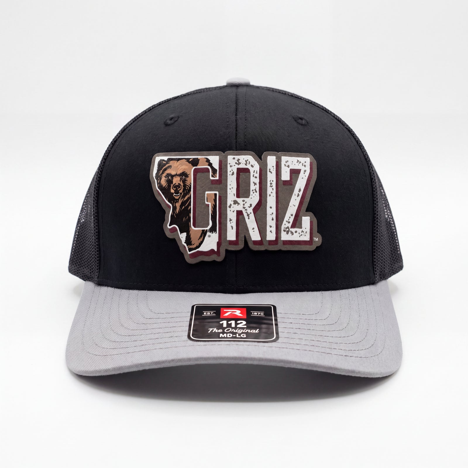 A black and grey Richardson 112 trucker hat with a printed leatherette patch of Griz with teh University of Montana Charging Bear in the G in the shape of Montana