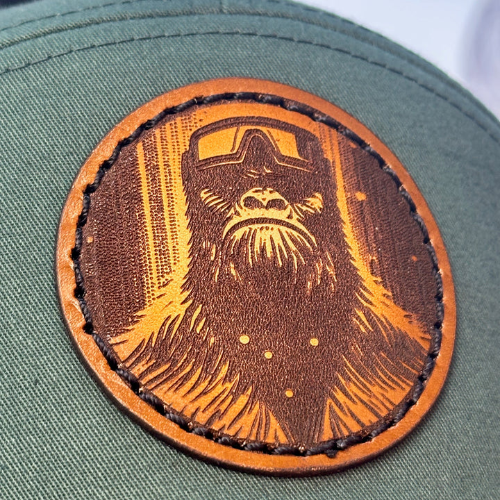 A close up of a hand dyed leather patch hat with bigfoot's face sewn onto a green Richardson flat bill trucker hat with black mesh.