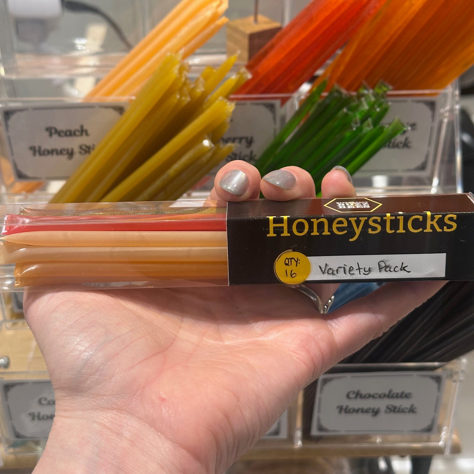 A hand holding a 16 pack of flavored honey sticks with colorful oney sticks in the background