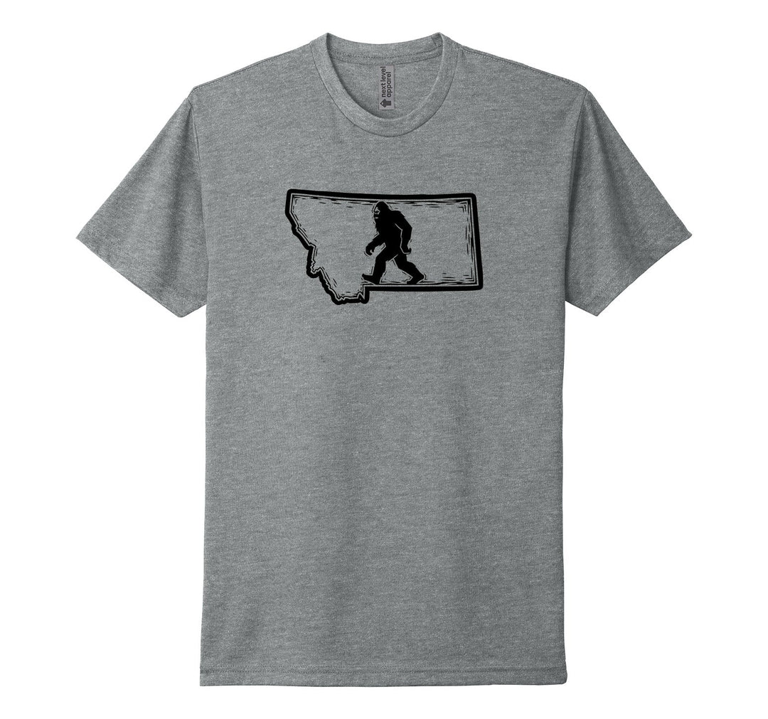 Dark heather grey Next Level Apparel unisex tee featuring a black woodcut-style illustration of Bigfoot striding through the outline of the state of Montana, designed and printed in Montana by Blue Peak Creative