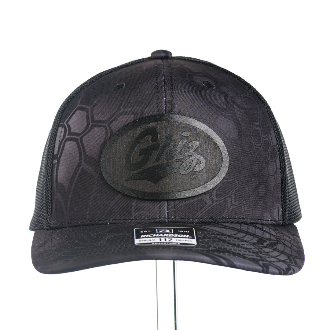 Front of a Black Leather Griz Script - Kryptek Trucker Cap—a black Kryptek camo Richardson 112 trucker hat with a black leather Griz Script patch, made in Missoula, Montana (home of the UM Grizzlies) by Blue Peak Creative