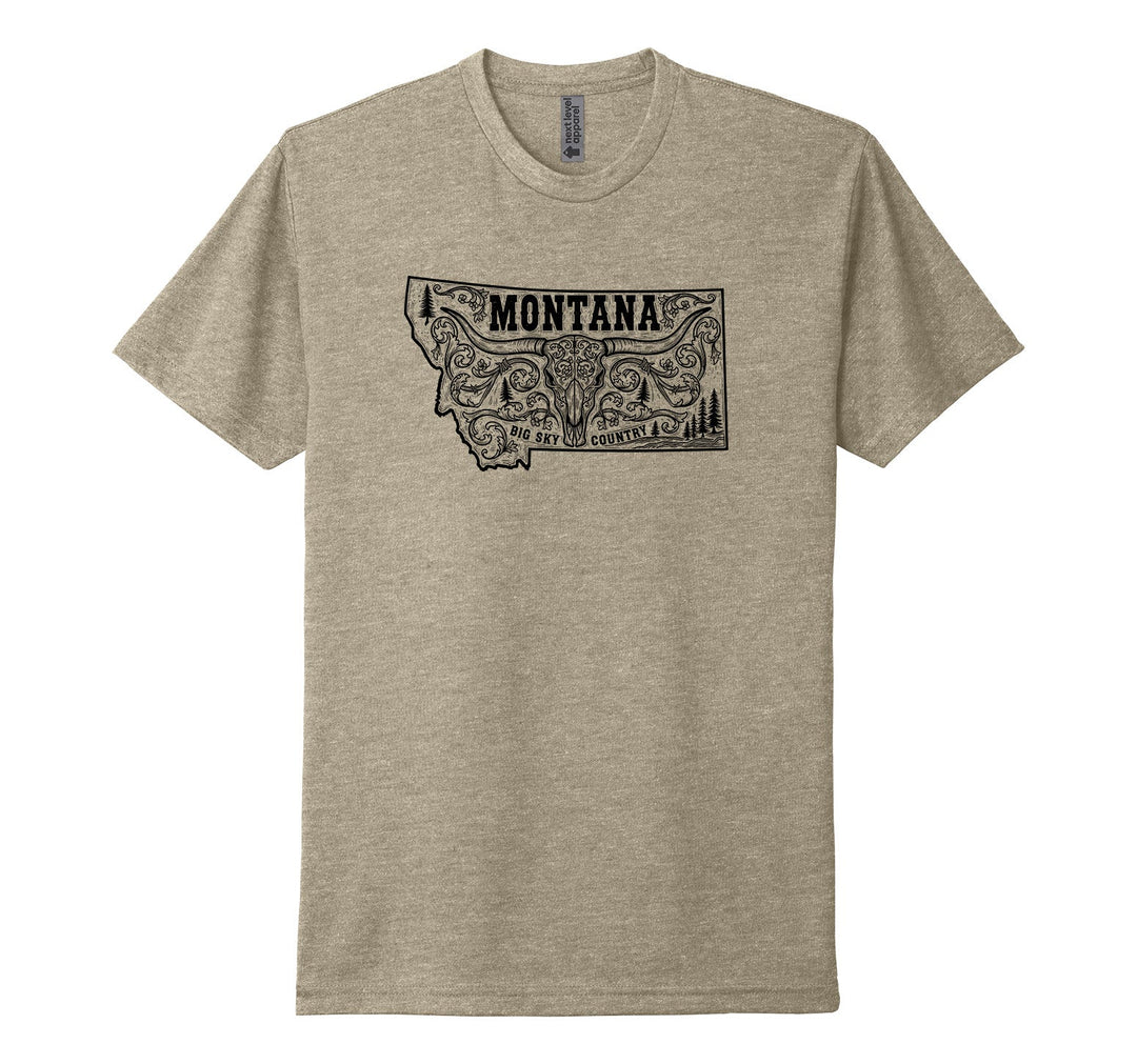 Heather tan Next Level Apparel unisex tee featuring a black illustration of an ornately decorated longhorn cow skull, surrounded by decorative flourishes and trees, beneath the word Montana, and above the text Big Sky Country—all within the outline of the state of Montana, designed and printed in Montana by Blue Peak Creative