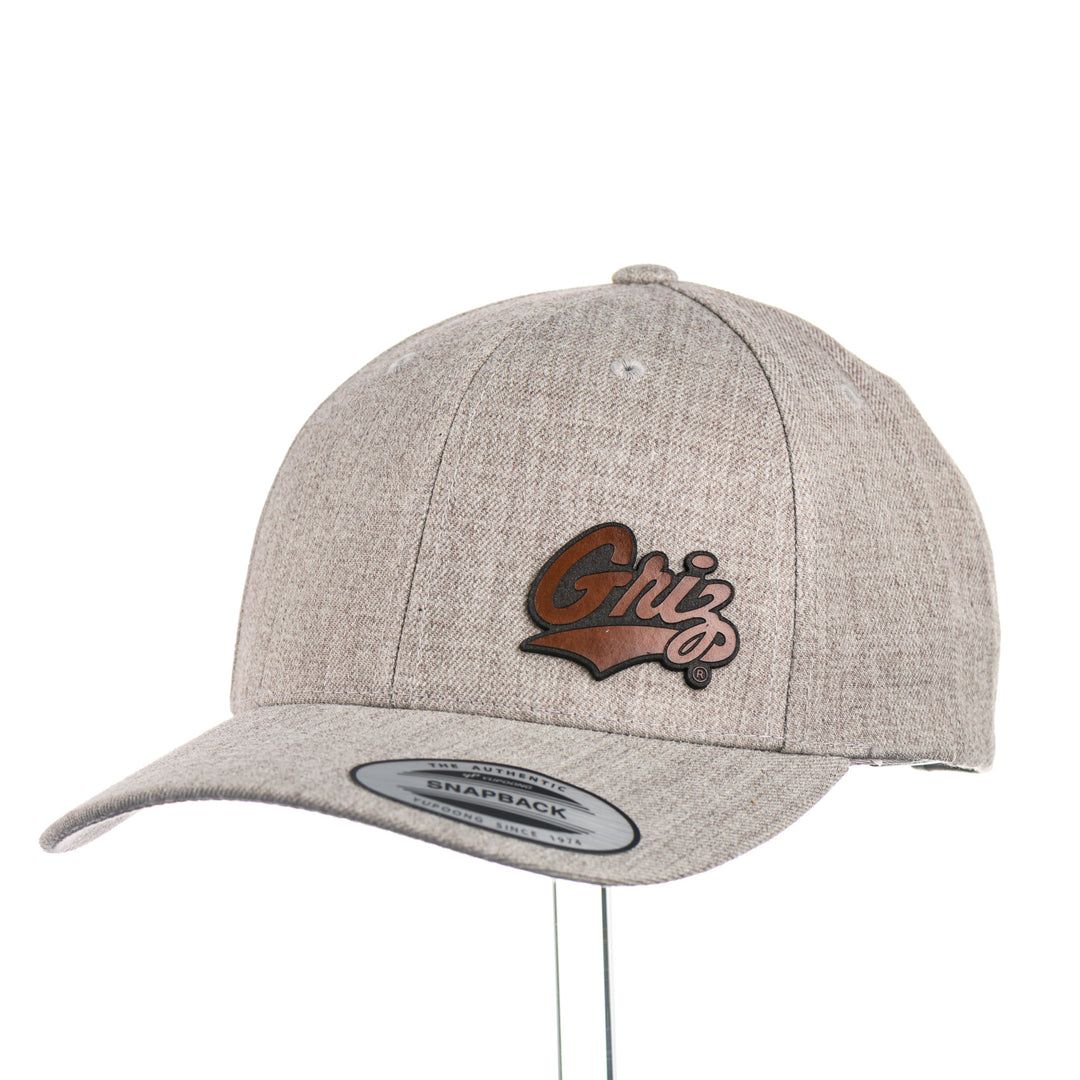 3/4 view of a Tiny Leather Griz Script - Premium Wool Blend Cap—a heather grey premium wool blend snapback hat with a small brown leather Griz Script patch, made in Missoula, Montana (home of the UM Grizzlies) by Blue Peak Creative