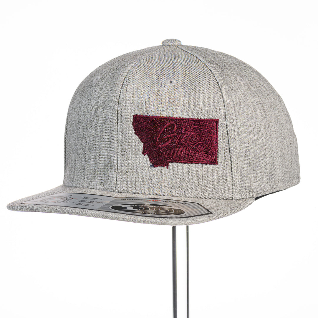 Grey Montana Griz Script - Flat Bill Hat made in Missoula, MT (home of the UM Grizzlies) by Blue Peak Creative—a grey flat bill hat embroidered with the University of Montana Griz script inside the outlined silhouette of the state of Montana (3/4 view).