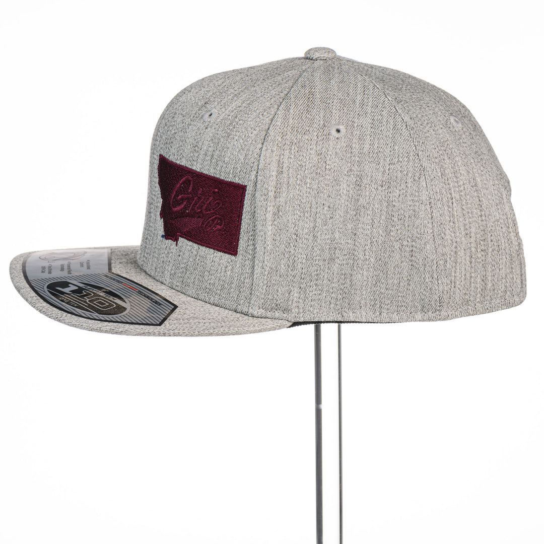 Grey Montana Griz Script - Flat Bill Hat made in Missoula, MT (home of the UM Grizzlies) by Blue Peak Creative—a grey flat bill hat embroidered with the University of Montana Griz script inside the outlined silhouette of the state of Montana (side).