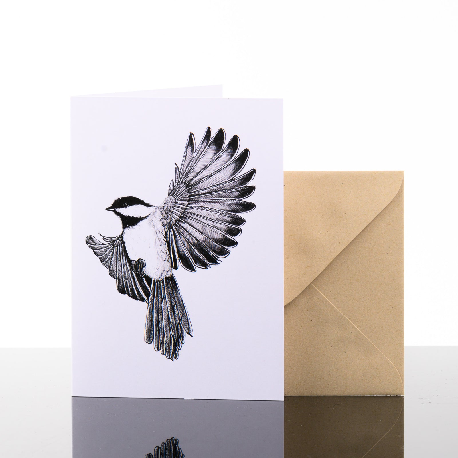 Blank greeting card featuring original black and white artwork by Brittany Finch - a Black-capped Chickadee, arrested mid-air with wings spread wide - and kraft brown envelope by Brittany Finch Art in Montana