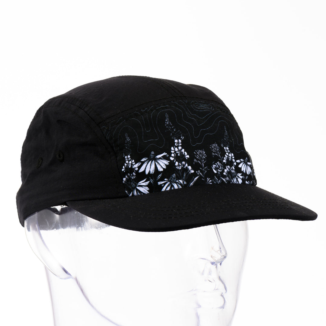 3/4 view of a black 5-panel hat with a brim featuring original black and white artwork by Brittany Finch -  wildflowers stretched across the front panel -  made in Montana by Brittany Finch Art and displayed on a mannequin head 