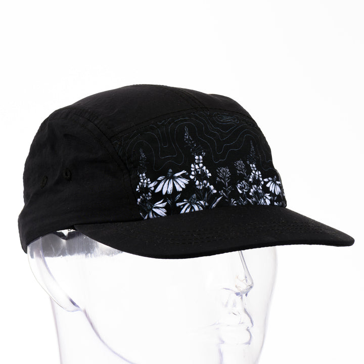 3/4 view of a black 5-panel hat with a brim featuring original black and white artwork by Brittany Finch -  wildflowers stretched across the front panel -  made in Montana by Brittany Finch Art and displayed on a mannequin head 