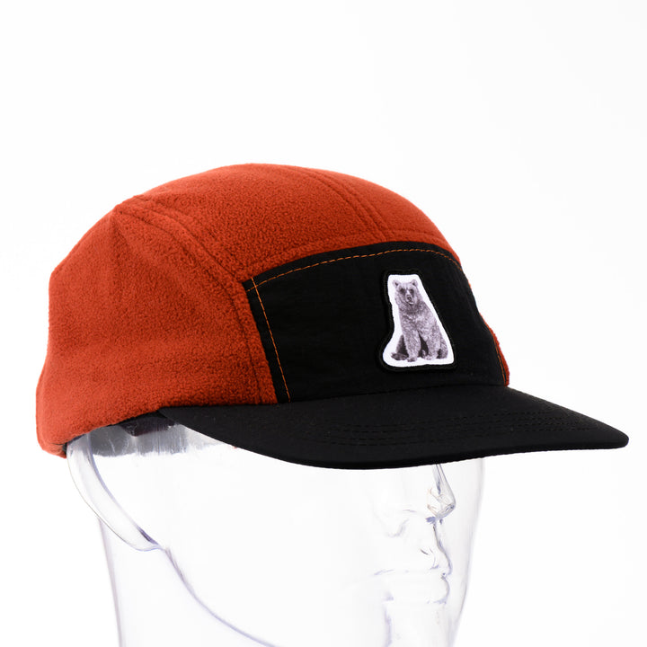 3/4 view of an orange and black fleece 5-panel hat with a brim and a patch featuring original black and white artwork by Brittany Finch -  a bear sitting on its haunches -  made in Montana by Brittany Finch Art and displayed on a mannequin head 