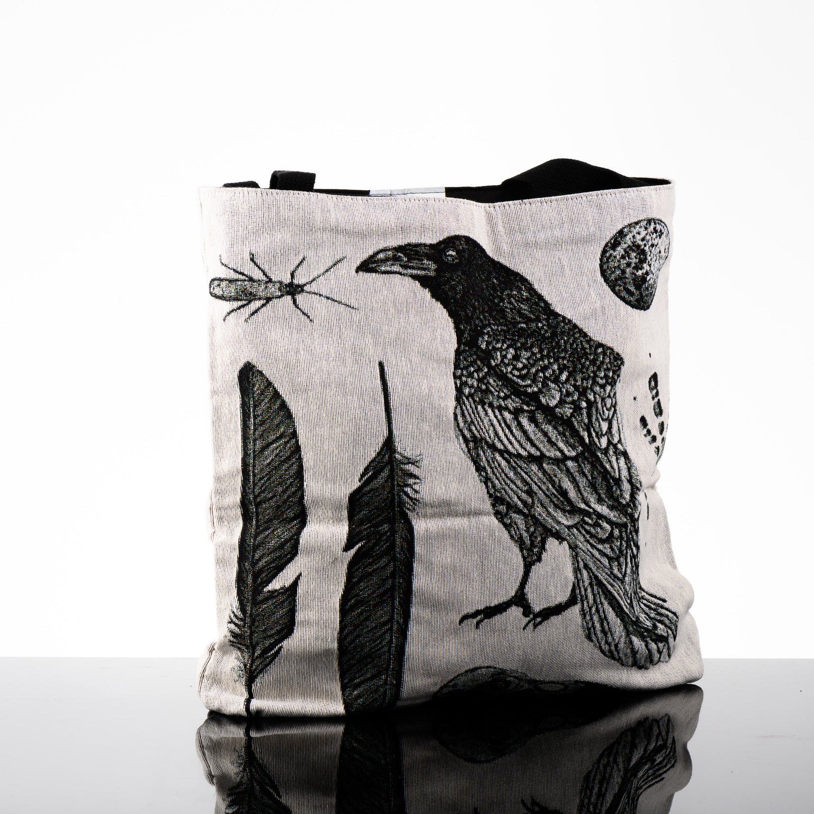 White woven tote bag featuring original black and white artwork by Brittany Finch - a study of a raven, two feathers, an insect, and the moon -  made in Montana by Brittany Finch Art