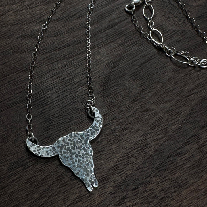 A sterling silver textured buffalo skull necklace with a silver chain on a dark wood background