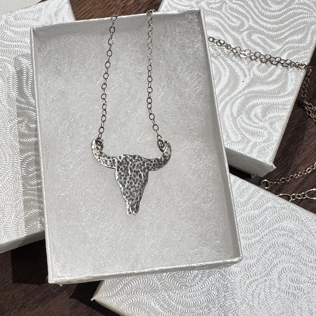 A sterling silver textured buffalo skull pendant necklace with a silver chain in a white jewelry box on a dark wood background.