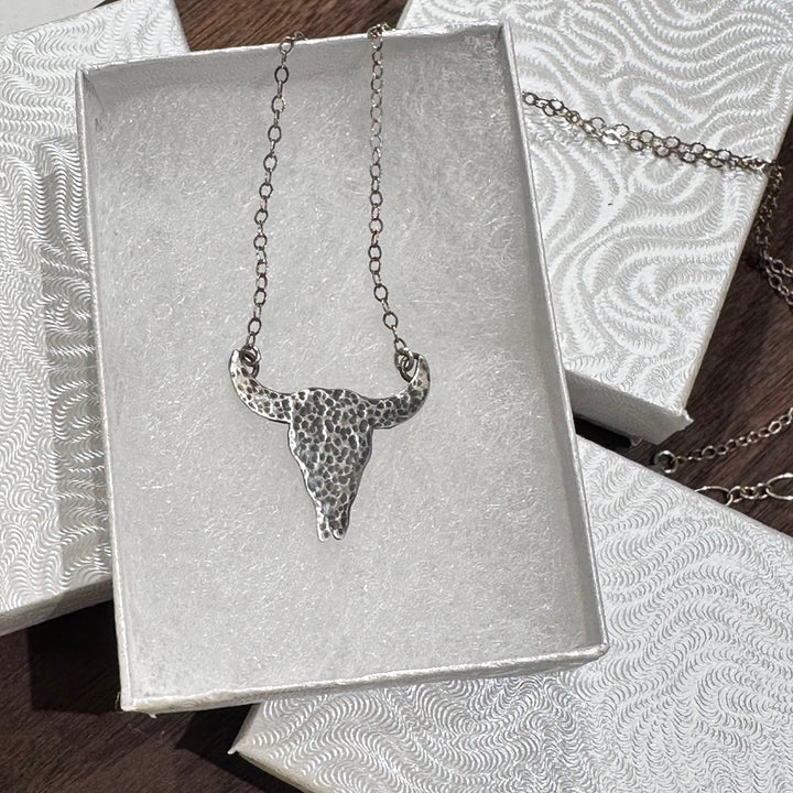 A sterling silver textured buffalo skull pendant necklace with a silver chain in a white jewelry box on a dark wood background.