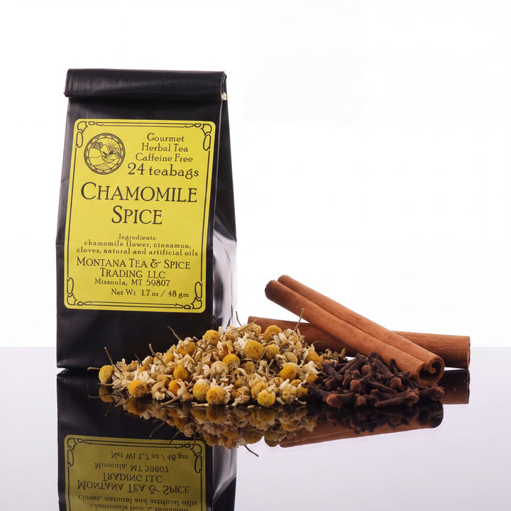 A black stand-up pouch of Chamomile Spice herbal tea with a yellow label, shown beside loose dried chamomile flowers, cinnamon sticks, and whole cloves on a reflective white surface.