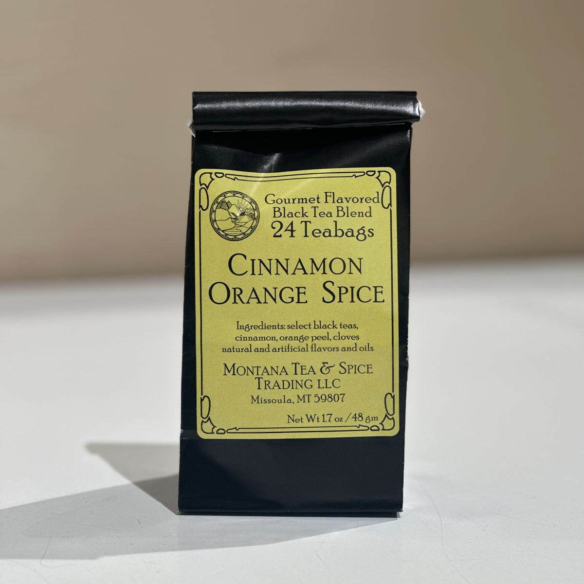 A black tea bag package labeled 'Cinnamon Orange Spice' by Montana Tea & Spice Trading LLC, photographed upright on a light surface against a plain beige background.