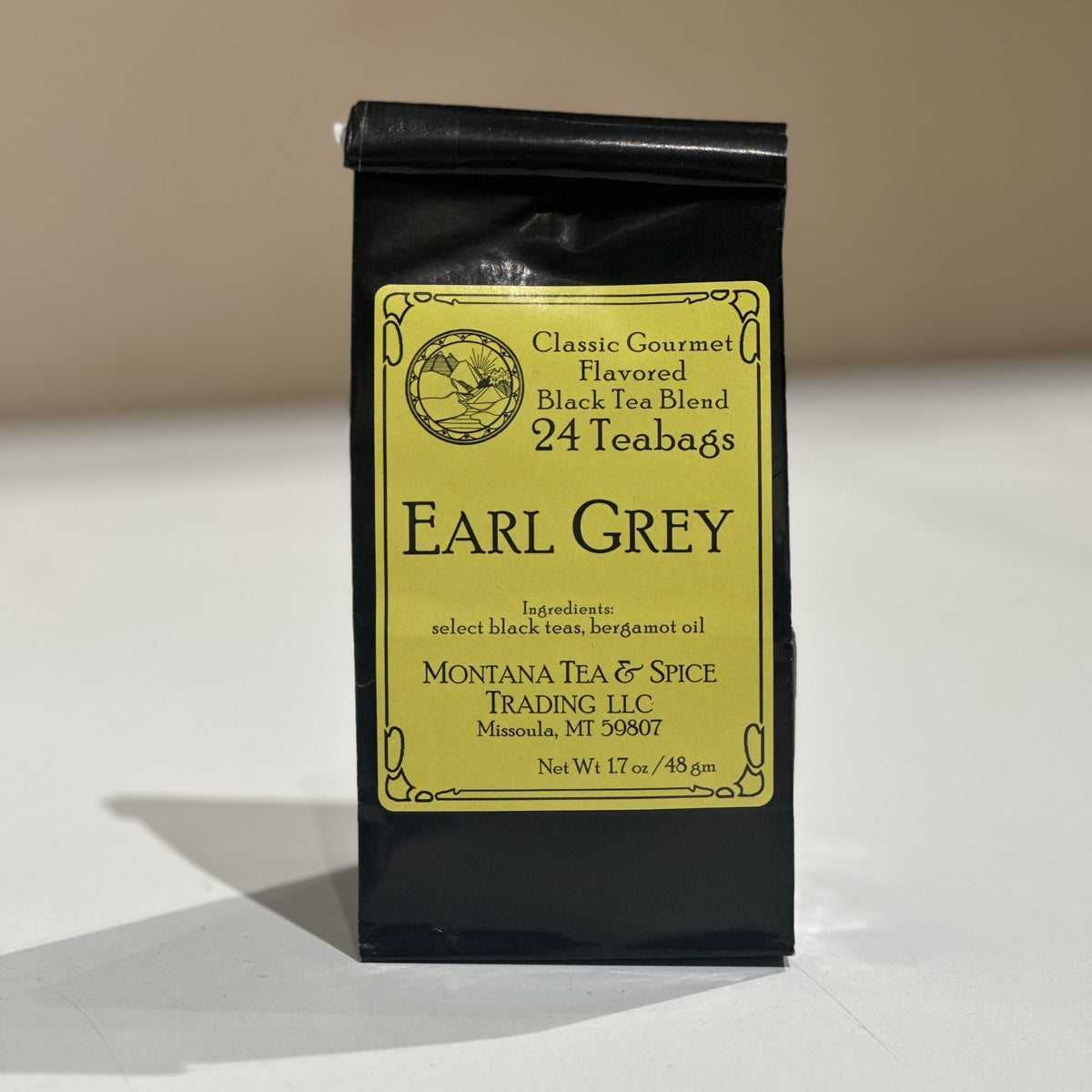 A black stand-up pouch of Earl Grey tea with a large yellow label, photographed on a light surface against a beige background. The label reads Classic Gourmet Flavored Black Tea Blend, 24 Teabags, and shows Montana Tea & Spice Trading LLC.
