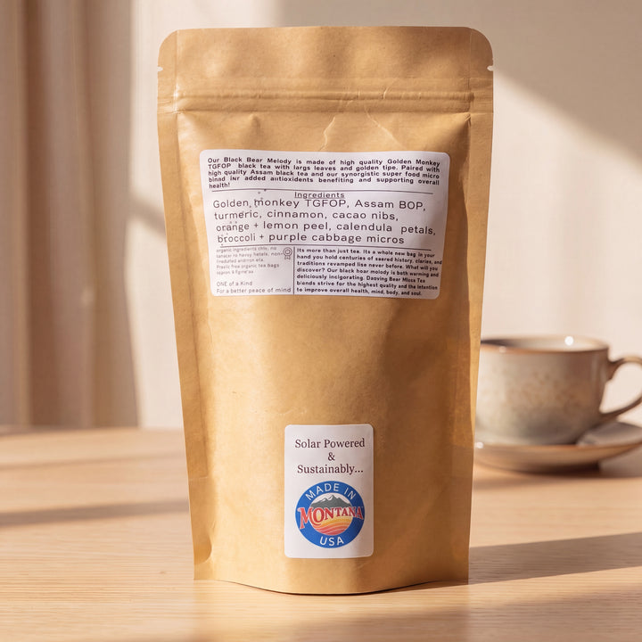 A kraft paper stand-up pouch of micro green tea stands on a wooden table in warm sunlight, with a ceramic cup on a saucer blurred in the background. The label reads Solar powered and sustainably Made in Montana