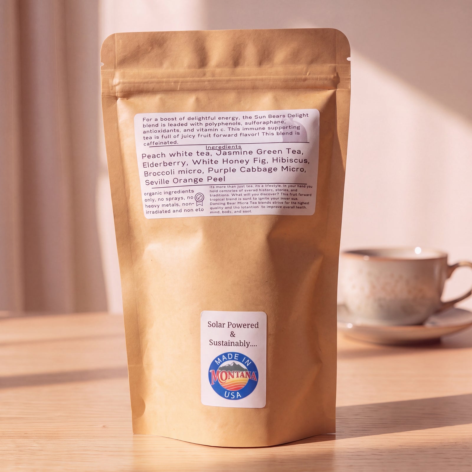 A kraft paper stand-up pouch of tea sits on a wooden surface with a blurred tea cup in the background. The pouch has a white ingredient label and a lower white sticker that reads 'Solar Powered & Sustainably Made in Montana'
