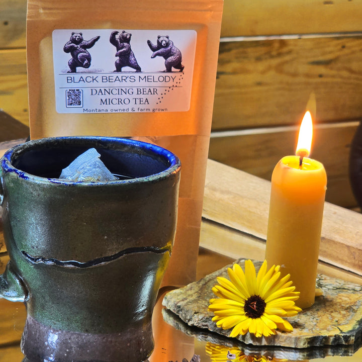 Tea package containing Black Bears Melody Micro Tea grown in Montana with a teabag, ceramic mug, candle, and yellow flower on a wooden surface.