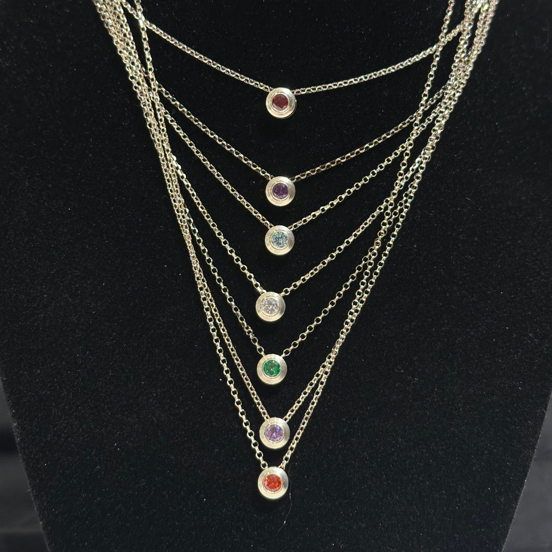 Seven necklaces with different colored cubic zirconia birthday stones inlayed on a black background
