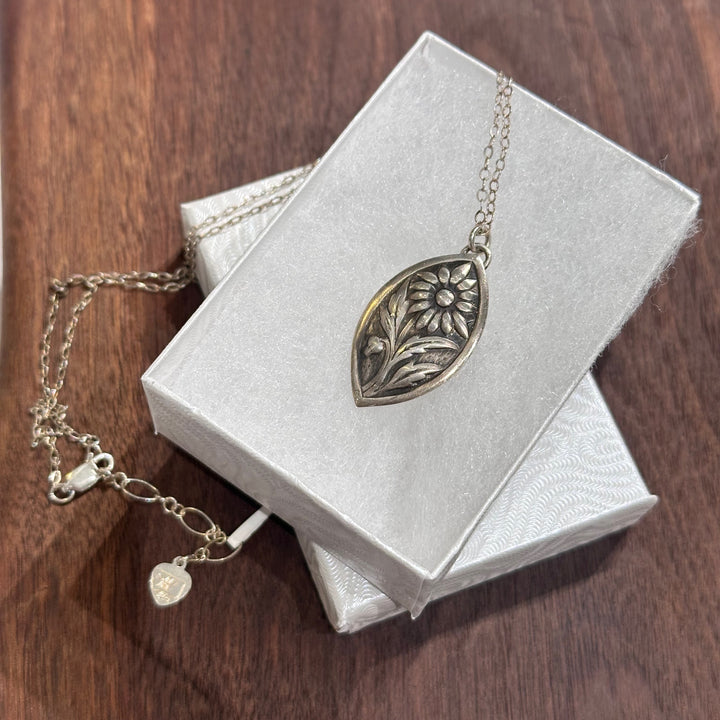 A sterling silver flower pendant necklace handmade by Cobblestone Designs of Montana in a white gift box on a dark wood background