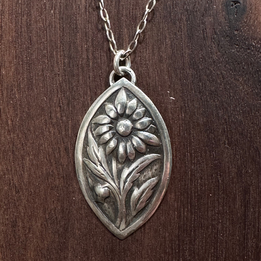 A close up of a sterling silver flower pendant necklace handmade by Cobblestone Designs of Montana on a dark wood background