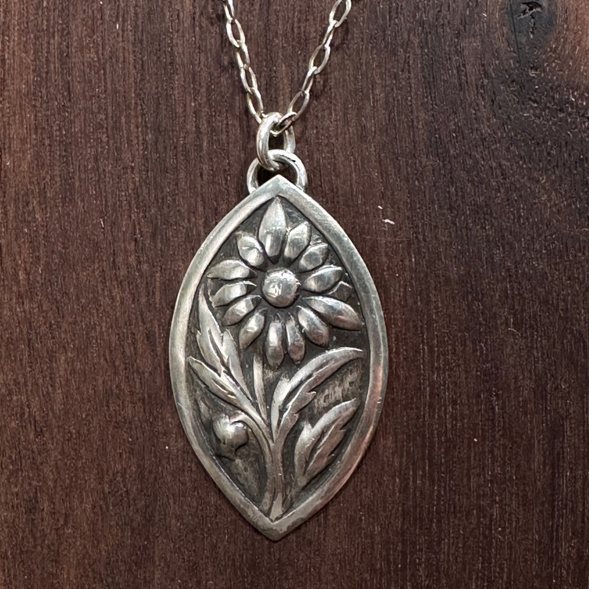 A close up of a sterling silver flower pendant necklace handmade by Cobblestone Designs of Montana on a dark wood background