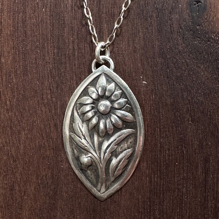 A close up of a sterling silver flower pendant necklace handmade by Cobblestone Designs of Montana on a dark wood background