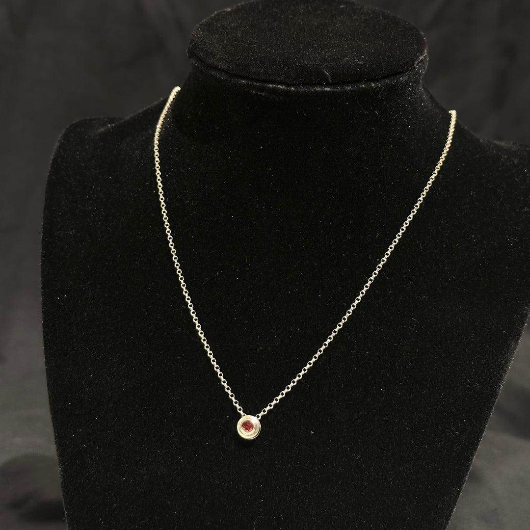 A garnet colored cubic zirconia in a round sterling silver bezel on a silver chain on a black background