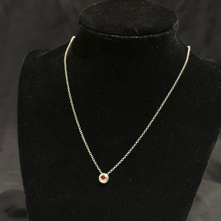 A garnet colored cubic zirconia in a round sterling silver bezel on a silver chain on a black background