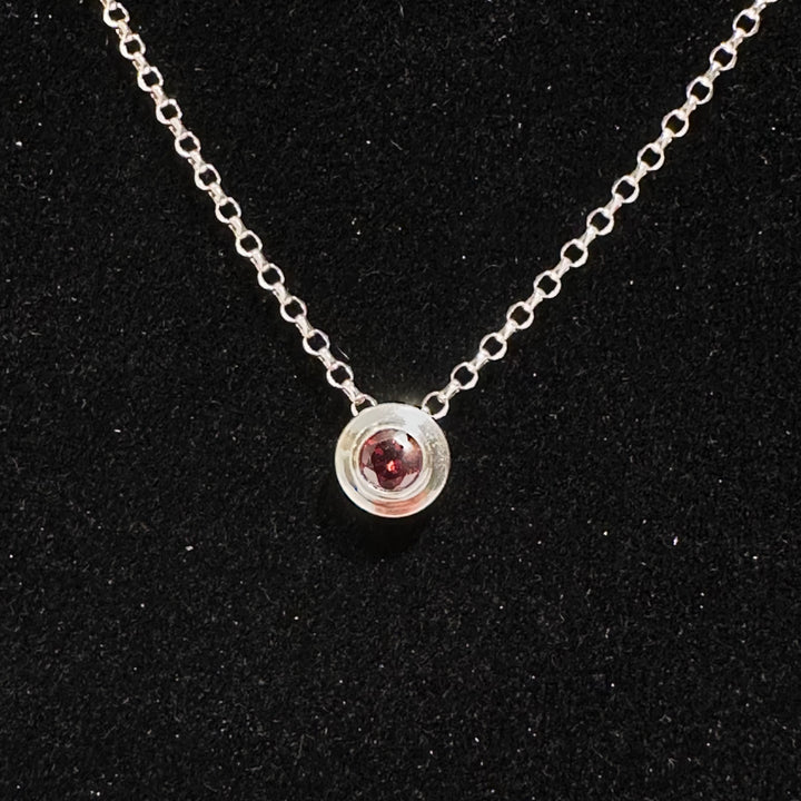 A close up of a January birthday necklace garnet colored cubic zirconia in a sterling silver bezel on a silver chain on a black background.