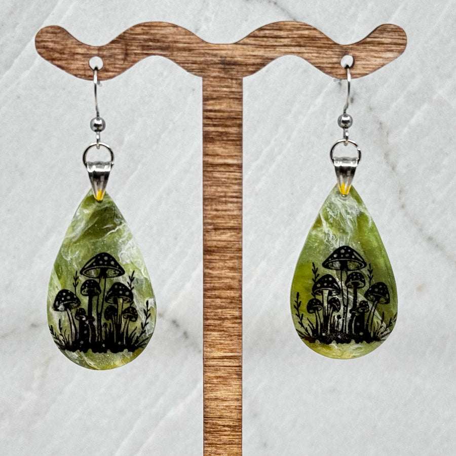 Pair of Large Teardrop Earrings with Mushrooms by Bitterroot Shining Creations, featuring a cluster of black mushrooms against a marbleized green polymer clay backdrop, hanging