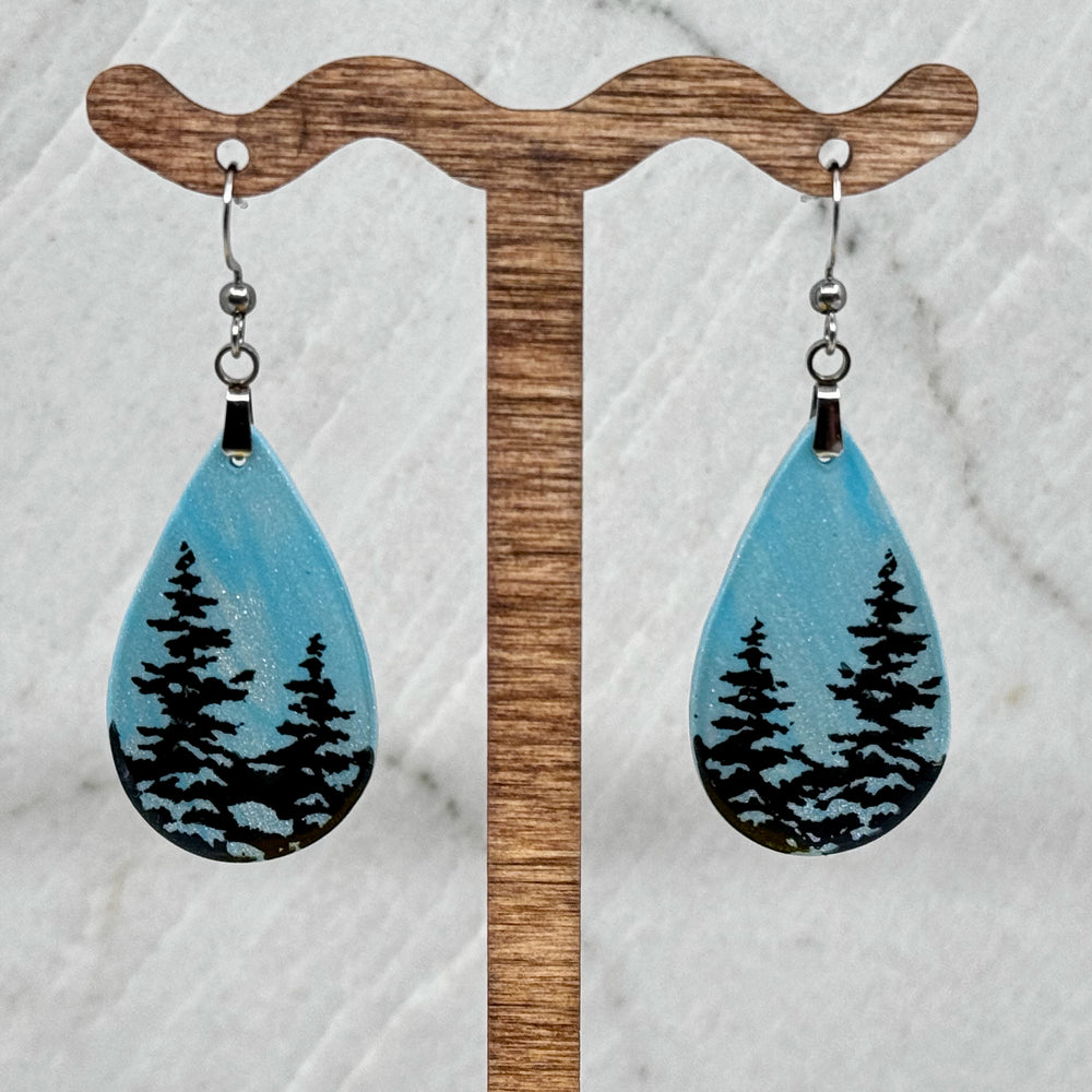 Pair of Large Teardrop Earrings with Trees, by Bitterroot Shining Creations, featuring tree silhouettes against a sparkly light blue polymer clay background (2 trees), hanging