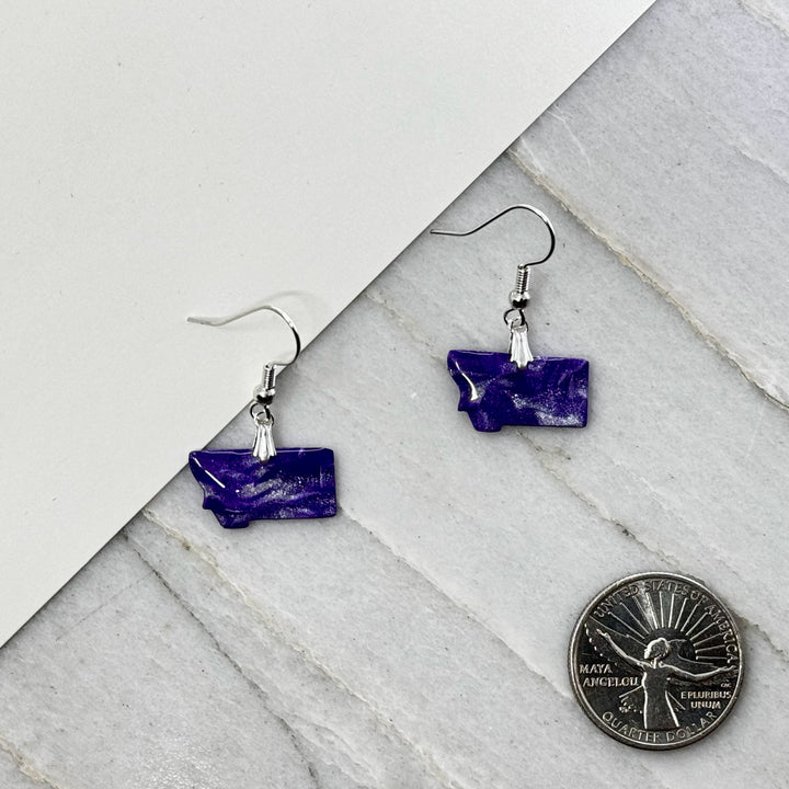 Pair of small, polymer clay Montana Earrings by Bitterroot Shining Creations in assorted sparkly colors (purple and white), with scale