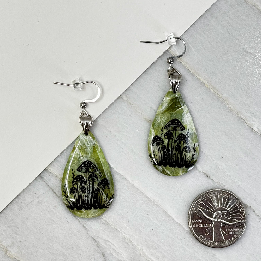 Pair of Large Teardrop Earrings with Mushrooms by Bitterroot Shining Creations, featuring a cluster of black mushrooms against a marbleized green polymer clay backdrop, with scale