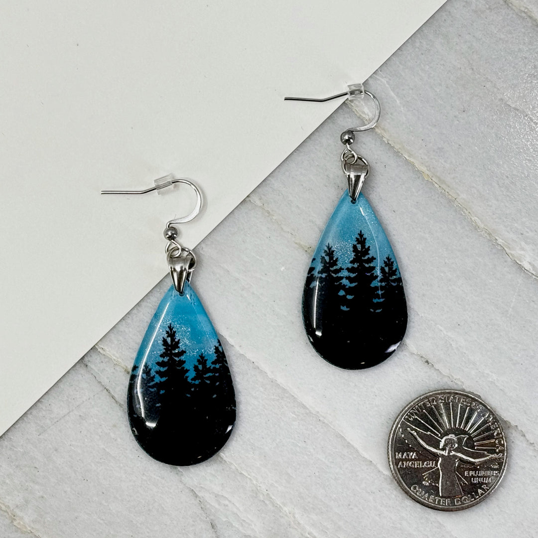 Pair of Large Teardrop Earrings with Trees, by Bitterroot Shining Creations, featuring tree silhouettes against a sparkly light blue polymer clay background (3-4 trees), with scale