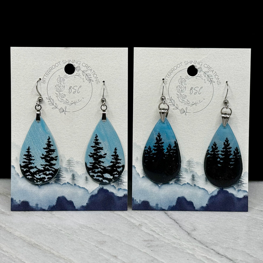 Two pairs of Large Teardrop Earrings with Trees, by Bitterroot Shining Creations, featuring tree silhouettes against a sparkly light blue polymer clay background, on cards