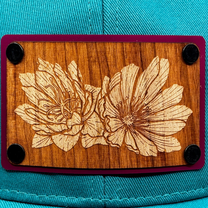 Last Best Supply Co's Bitterroot Cherry & Pink Metal Patch on Evergreen Eco Trucker Hat, featuring a cherry wood patch engraved with bitterroot flowers (patch detail)